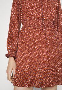 Κόκκινο φόρεμα με σχέδιο και μαζεμένη μέση και μακριά φουσκωτά μανίκια. Το ύφασμα διαθέτει λουλούδια με εναλλασσόμενα χρώματα και σχήματα.