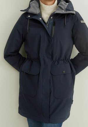 Osoba ubrana w granatową zimową kurtkę z kapturem, zamkiem błyskawicznym i kieszeniami na zatrzaski, z rękami w kieszeniach, w kremowym golfie.