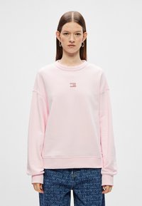 Jasnoróżowa bluza z okrągłym dekoltem i obniżonymi ramionami. Zawiera małe logo na środku. Miękka tkanina o gładkiej teksturze.