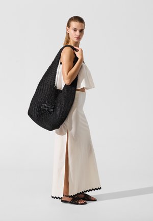 Sac tote noir tissé avec une finition texturée, associé à un haut blanc court et une longue jupe blanche avec une fente latérale et un ourlet festonné.