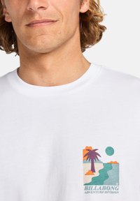 Fehér pamut póló, színes grafikával, amelyen pálmafa, folyó és nap látható, alján a "BILLABONG ADVENTURE DIVISION" felirattal.