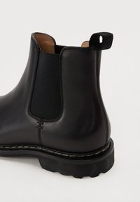 Musta nahkainen Chelsea-boot, jossa on joustavat sivupaneelit, pyöreä kärki, vetonauha ja kontrastiväri valkoinen ommel kumipohjassa.