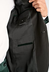 Hand holding open a black suit jacket showing inner label "JUNK de LUXE" and inner pockets against white shirt and green trousers.