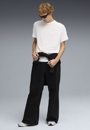 Ragazzo con occhiali da sole neri, t-shirt bianca Puma, pantaloni neri a gamba larga, sneakers bianche, che annoda una felpa nera intorno alla vita.