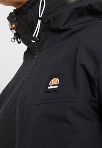 Svart huvtröja med dragkedja, snöre och en logotypapplikation i orange och rött. Materialet ser något strukturerat och lätt ut.