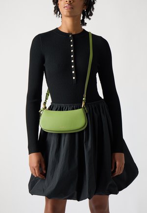 Groene leren crossbodytas met een gladde textuur, gebogen vorm en gouden hardware, gedragen over een zwarte geribbelde jurk en een volumineuze rok.