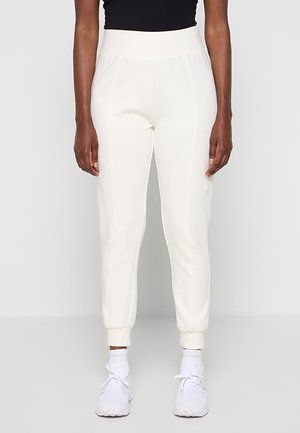 Personne portant un pantalon de jogging blanc taille haute, associé à des chaussettes blanches et des chaussures de sport blanches, debout devant un fond uni.