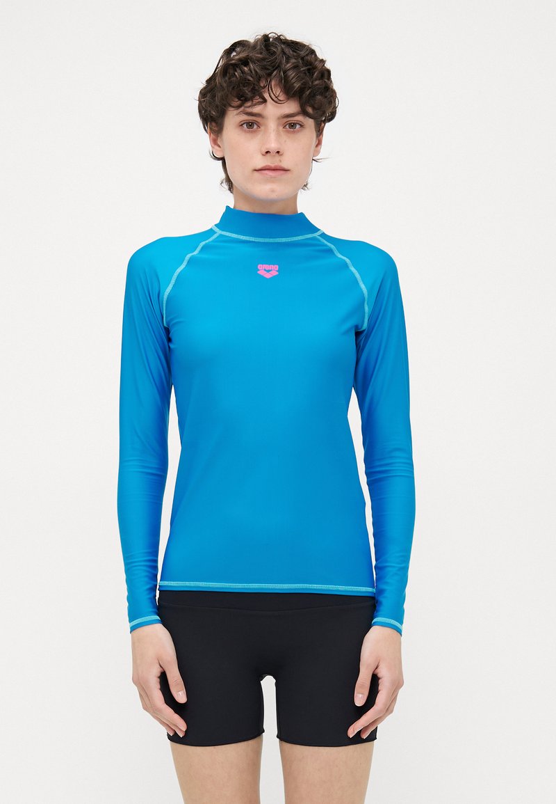 Camisa de rash guard de manga longa azul com gola alta e logótipo rosa. Apresenta costura de contraste e tecido leve e suave. Usada com calções pretos.