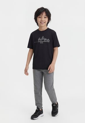 Ragazzo sorridente, indossa una maglietta nera con grafica, pantaloni della tuta grigi e scarpe da ginnastica nere, in piedi su uno sfondo bianco uniforme.