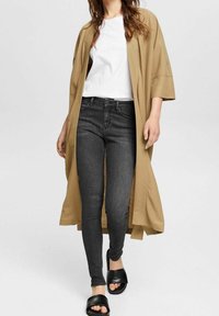 Cárdigan largo beige sobre una camiseta blanca, combinado con jeans ajustados gris oscuro y sandalias de deslizar negras. Texturas suaves, ajuste relajado.