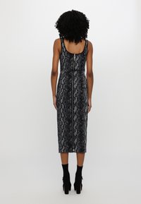 Femme aux cheveux bouclés portant une robe midi sans manches noire et grise à imprimé serpent avec une fermeture éclair au dos, associée à des bottines noires.