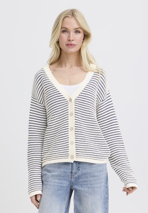 Blonde vrouw die een wit met marineblauw gestreepte cardigan met knopen draagt over een wit shirt en lichtblauwe spijkerbroek, staand tegen een effen achtergrond.