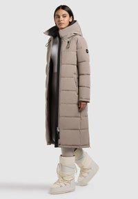 Lange, beige gewatteerde jas met een hoge kraag, zwarte accenten, zijritsen en een strakke afwerking. Gecombineerd met crèmekleurige winterlaarzen.