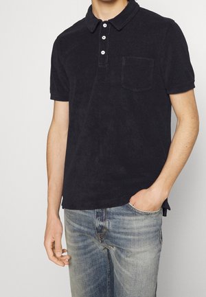 Homme portant un polo noir à manches courtes avec trois boutons blancs et une poche avant, associé à un jean bleu délavé.