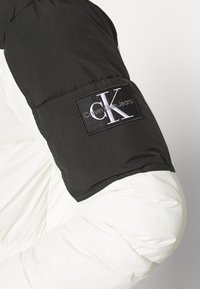 Rękaw czarno-białej kurtki puchowej z czarną naszywką z białym logo "cK Calvin Klein Jeans".