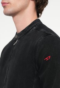 Melnas džinsa jaka ar apaļu apkakli, priekšējo rāvējslēdzēju un sarkanu izšuvuma akcentu uz kreisā pleca. Ar fakturētu virsmu.