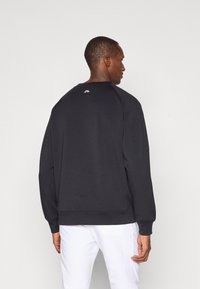 Fekete hosszú ujjú pulóver, kerek nyakkivágással és bordás mandzsettákkal. A hát felső részén egy kis logó található. Sima, pamutkeverék textúra.