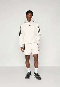 Vīrietis stāv bālā fona priekšā, tērpies baltā Adidas kapučjaka un šortos, ar melniem un neona akcentētiem sporta apaviem.