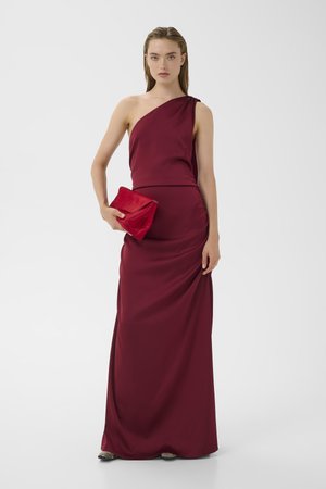 Woman in a floor-length, one-shoulder burgundy gown holding a red clutch bag against a plain white background.