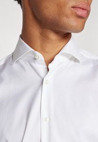 Camisa blanca de botones con un cuello clásico, que cuenta con dos botones en el cuello. La tela parece suave y ligeramente estructurada.