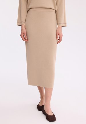 Beżowa ołówkowa spódnica do kolan noszona z dopasowanym topem i ciemnobrązowymi plecionymi balerinami na białym tle.