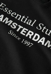 Hoodie noir avec un texte argenté inscrivant "Essential Studio AMSTERDAM Since 1997." Tissu lisse, coupe classique, sans motifs ni accents visibles.