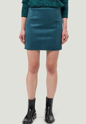 Mini-jupe en daim bleu sarcelle portée avec des bottines noires et un pull vert foncé tacheté, montrée de la taille aux pieds sur un fond neutre.