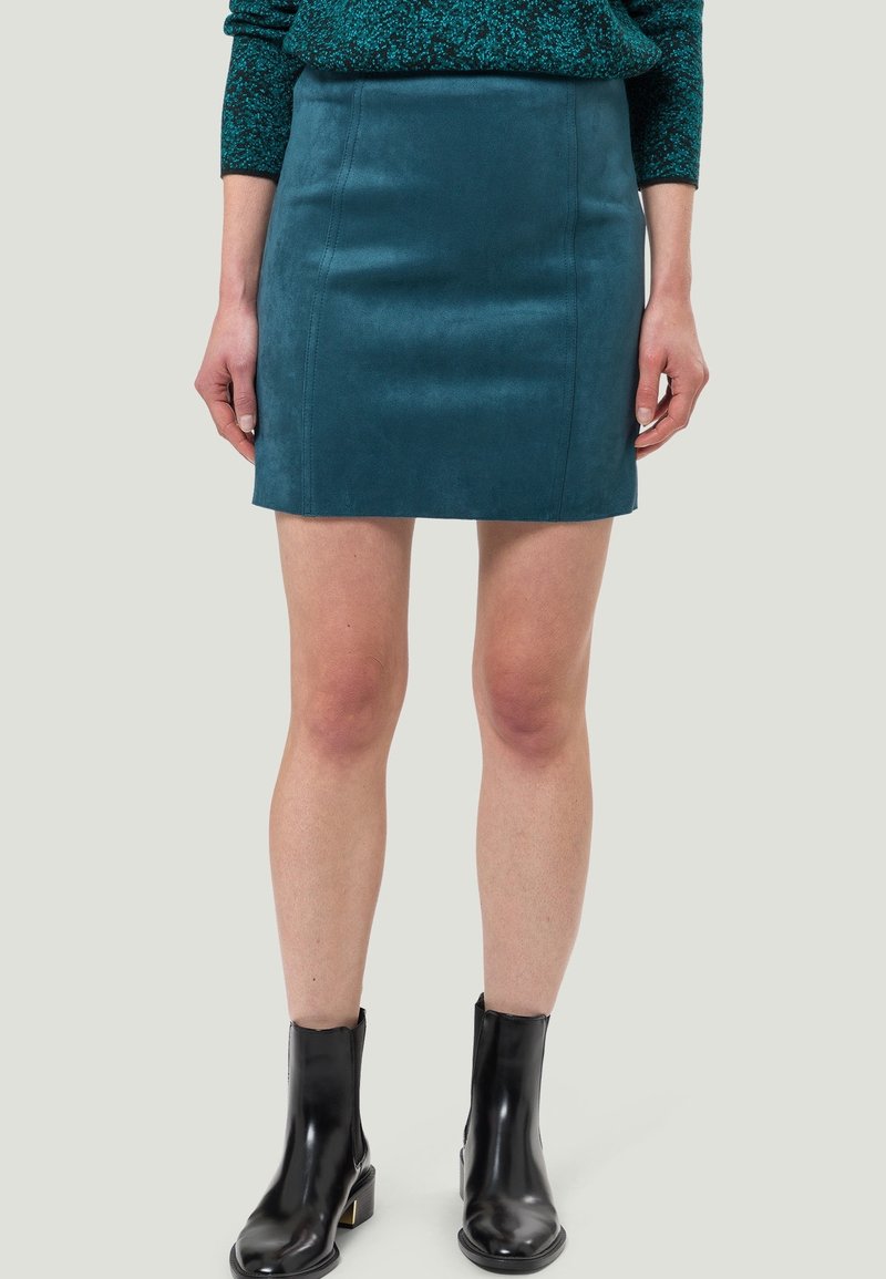 Mini-jupe en daim bleu sarcelle portée avec des bottines noires et un pull vert foncé tacheté, montrée de la taille aux pieds sur un fond neutre.