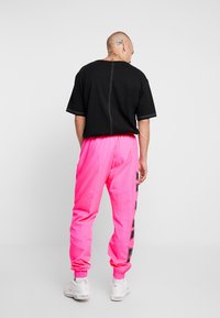 Man with tattoos wears black oversized T-shirt, bright pink joggers with black side text, and white sneakers, standing against plain light background.