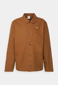 Brown cotton jacket with a button placket, two front pockets, a collar, and a small white Nike logo on the left chest.