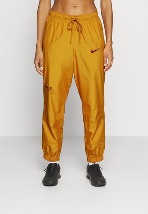 Persoon draagt mosterdgele Nike Paris team trainingsbroek en zwarte sneakers tegen een effen witte achtergrond.