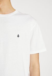 Witte katoenen t-shirt met een ronde hals, voorzien van een klein zwart geborduurd logo op de borst en korte mouwen.