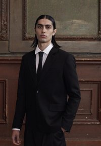 Black suit with a single button jacket and notch lapels, paired with a white shirt and black tie. Long dark hair, neutral background.