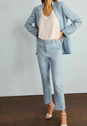 Kvinde poserende med hænderne på hofterne iført lyseblå blazer og bukse sæt, hvid ærmeløs top og hvide højhælede sandaler.