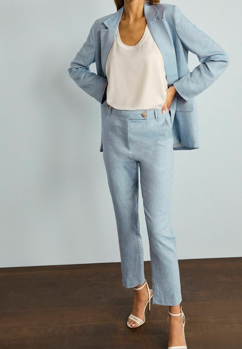 Femme posant les mains sur les hanches, portant un ensemble blazer et pantalon bleu clair, un haut sans manches blanc et des sandales blanches à talons hauts.