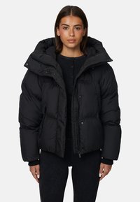 Giacca a vento nera con cappuccio oversize, texture trapuntata, chiusure con zip e bottoni a pressione, e tasche laterali. Indossata sopra un top nero.