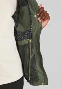 Main tenant une veste vert olive ouverte montrant une poche intérieure, une doublure à motifs et un détail de cordon sur un fond neutre.