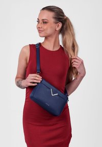 Námořnická křížová kabelka vyrobená z hladké kůže se zlatým V-úhlovým akcentem a nastavovacími popruhy, nošená přes červené šaty.