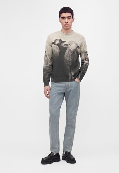 Homme portant un pull avec un grand motif noir et blanc de moutons, un jean bleu clair et des chaussures noires, debout devant un fond clair uni.