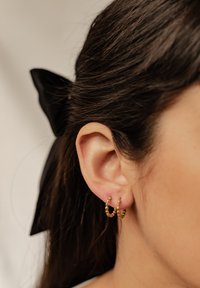 Złote kolczyki w kształcie obręczy z teksturowanym wzorem i małymi koralikami. Kolczyki noszone są w przekutym uchu, częściowo widoczne ciemne włosy.