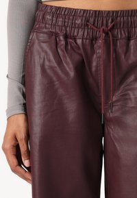 Burgundske hlače iz umetnega usnja z elastičnim pasom, zavezovanjem in gladko teksturo, delno prikazane z roko, ki počiva na tkanini.