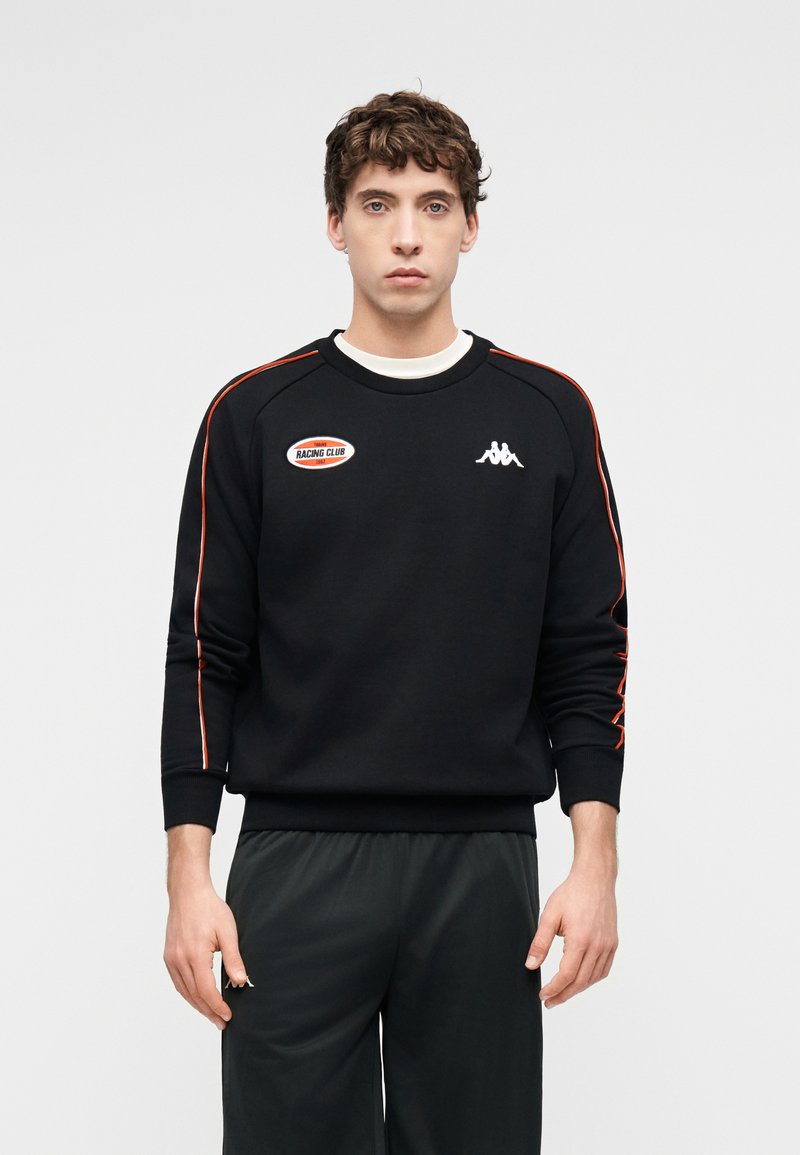 Νέος άνδρας με κοντά σγουρά μαλλιά που φοράει μαύρο φούτερ με κόκκινες ρίγες και λογότυπα, στέκεται μπροστά σε λευκό απλό φόντο.