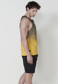Rumena in črna gradient majica brez rokavov z ovalnim izrezom in širokimi armholes; v kombinaciji s črnimi tovornimi kratkimi hlačami s stranskimi žepi.