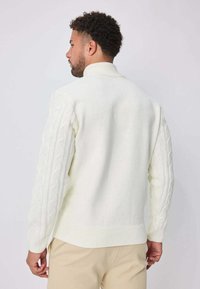 Pull en maille blanche avec un col haut et des manches à motif torsadé. Comprend des poignets et une taille côtelés, une texture lisse et une coupe décontractée.