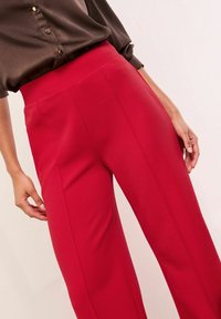 Piros, magas derekú nadrág sima, texturált anyagból. Testhez álló fazonnal és oldalsó zsebekkel rendelkezik. Sötét gombos blúzzal párosítva.