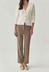 Cárdigan blanco acanalado con escote en V y botones de madera, combinado con pantalones marrones a medida y zapatos de tela beige. Texturas suaves en todo.