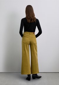 Pantalones de pierna ancha en color mostaza, de talle alto, con bolsillos traseros y una etiqueta de cuero en la parte trasera; combinados con una camiseta negra de manga larga.