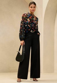 Blusa nera a fiori con maniche lunghe, abbinata a pantaloni a gamba larga neri a vita alta e una borsa nera. Tessuto morbido, leggero luccichio.