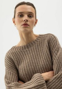 Junge Frau mit heller Haut und braunem, nach hinten gebundenem Haar, die kleine Creolenohrringe und einen grob gestrickten taupefarbenen Pullover trägt und die Arme verschränkt hat.