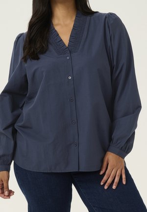 Kobieta ubrana w granatową bluzkę z długim rękawem zapinaną na guziki z marszczeniami przy dekolcie w kształcie litery V oraz ciemnoniebieskie jeansy, na jednolitym tle.